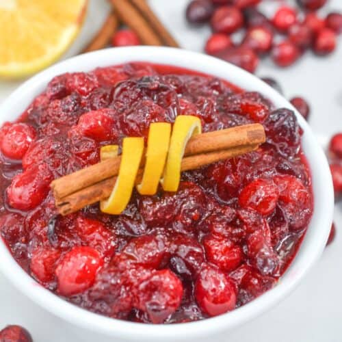 A bowl of Orange Cranberry Sauce garnished with a cinnamon stick and lemon peel, with fresh cranberries, cinnamon sticks, and a lemon slice in the background.