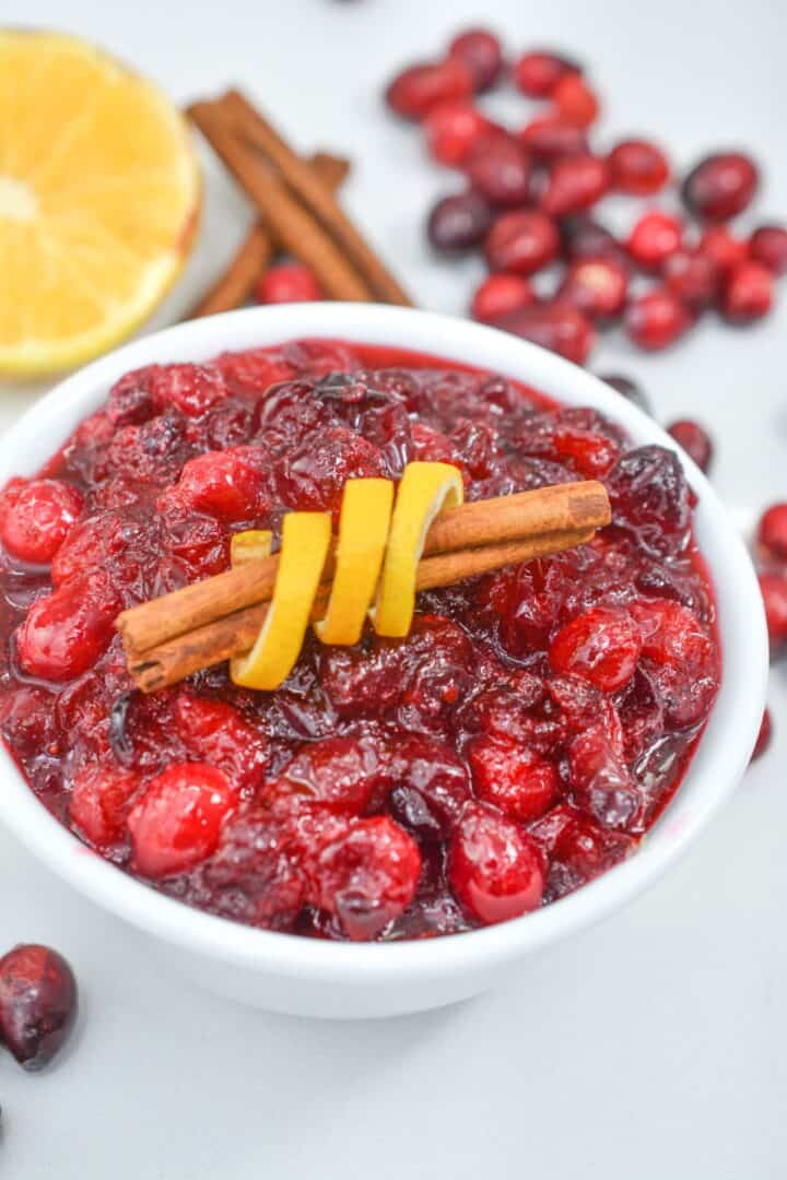 A bowl of Orange Cranberry Sauce garnished with a cinnamon stick and lemon peel, with fresh cranberries, cinnamon sticks, and a lemon slice in the background.