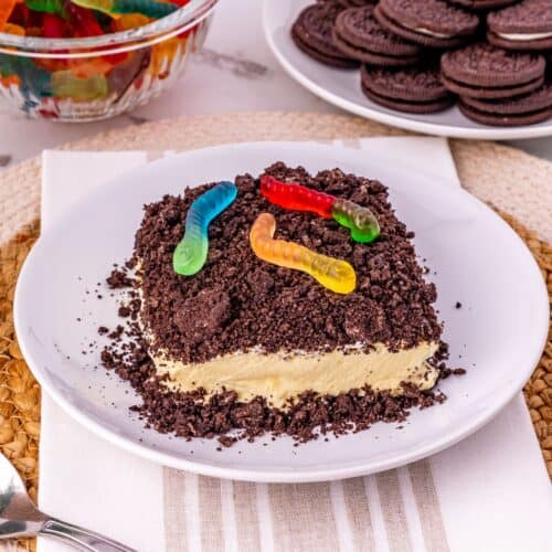 An Oreo Dirt Cake with crumbled cookies and gummy worms on top sits on a plate; nearby are a bowl of gummy worms and a plate of chocolate sandwich cookies.