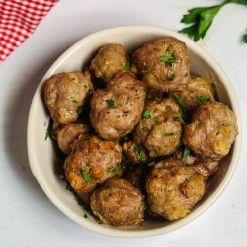 A bowl of baked meatballs garnished with chopped herbs sits on a white surface, accompanied by a red checked cloth and vibrant green leafy vegetables.