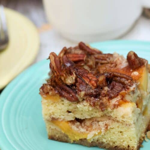 A square slice of peach coffee cake topped with pecans sits on a light blue plate, with a cup of coffee in the background.
