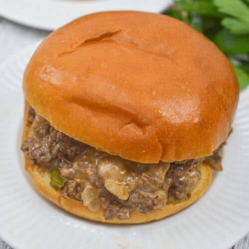 A Philly Cheese Steak Sloppy Joe rests on a white plate, crowned with a glossy bun and packed with seasoned ground beef and onions. In the background, fresh parsley adds a pop of color.