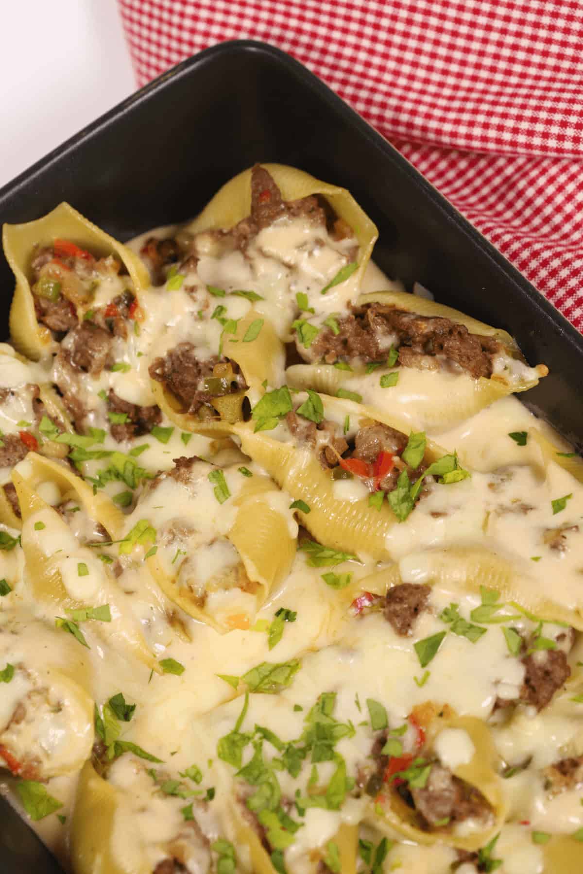 Philly Cheesesteak Stuffed Shells with ground meat and melted cheese, garnished with fresh parsley, in a black baking dish on a red checkered cloth.