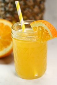 A mason jar filled with orange juice, garnished with an orange slice and a yellow striped straw, evokes the vibrant flavor of summer. In the blurred background, a whole orange rests alongside apple pie moonshine to inspire delicious seasonal indulgence.