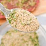 A close-up of a spoon holding a mixed dry herb and spice Pot Roast Seasoning recipe, with a bowl of the same mixture and a piece of meat in the background.