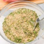 A glass bowl filled with a dry herb and spice mixture for a classic Pot Roast Seasoning recipe sits next to a piece of raw meat coated with the flavorful blend.