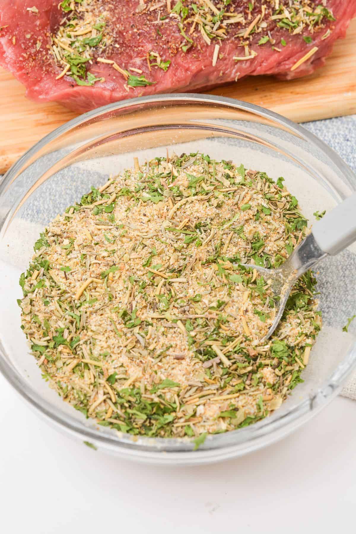 A glass bowl filled with a dry herb and spice mixture for a classic Pot Roast Seasoning recipe sits next to a piece of raw meat coated with the flavorful blend.
