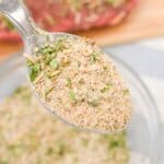 A close-up of a spoon holding the recipe blend of dried herbs and spices, with a glass bowl of the mixture and a piece of raw meat in the background.