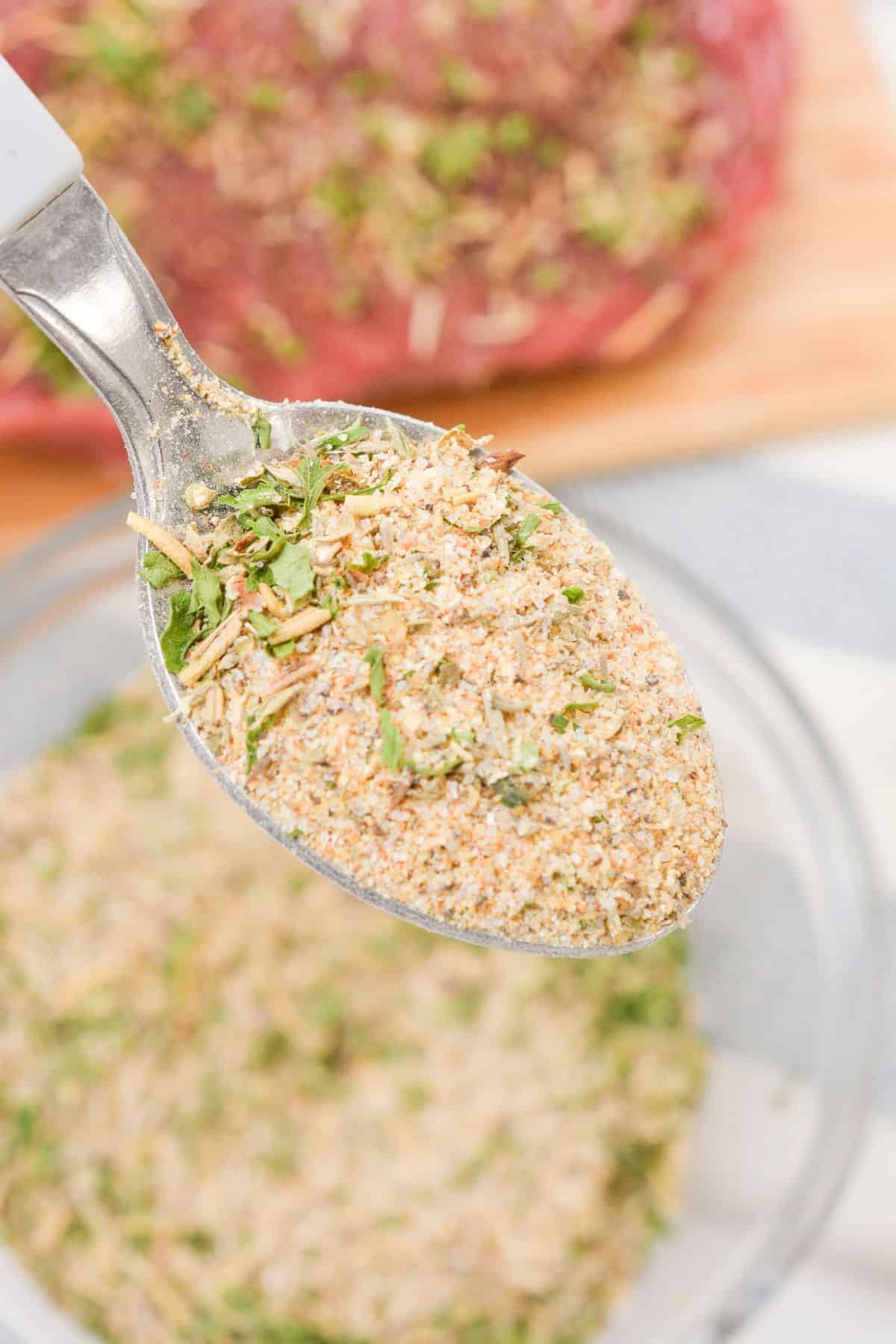 A close-up of a spoon holding  the recipe blend of dried herbs and spices, with a glass bowl of the mixture and a piece of raw meat in the background.