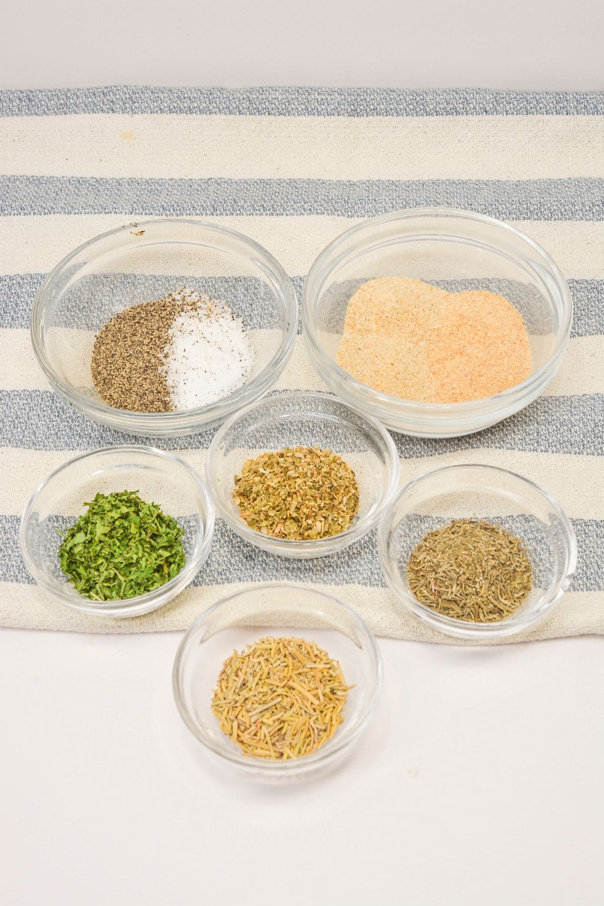 Seven small glass bowls filled with various dried herbs and spices, perfect for creating a recipe, are arranged on a striped cloth background.