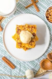 A square slice of Pumpkin Crunch Cake topped with ice cream sits on a white plate, surrounded by pecans, cinnamon sticks, and a glass of milk for the perfect autumn treat.