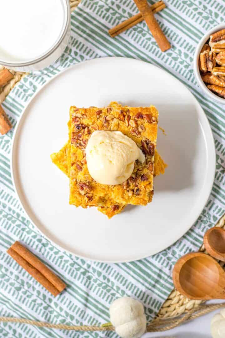 A square slice of Pumpkin Crunch Cake topped with ice cream sits on a white plate, surrounded by pecans, cinnamon sticks, and a glass of milk for the perfect autumn treat.