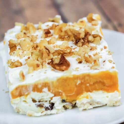 A square slice of Lush Cake with cream and orange filling, topped with chopped walnuts, served on a white plate.