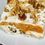 A slice of Lush Cake with a creamy white base, orange filling, whipped topping, and chopped walnuts is served on a white plate.