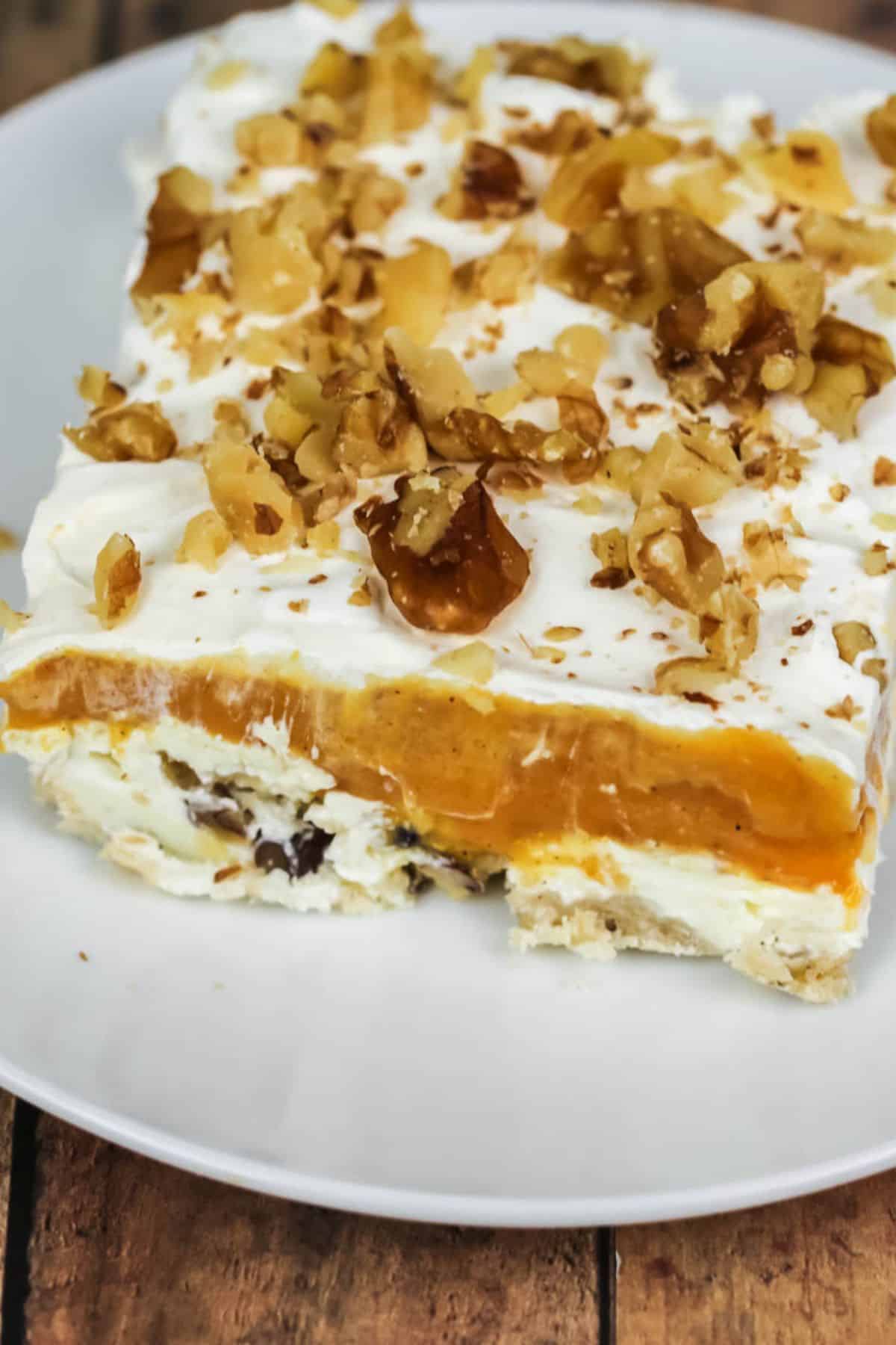 A slice of Lush Cake with a creamy white base, orange filling, whipped topping, and chopped walnuts is served on a white plate.