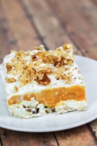 A square slice with a creamy white base, orange middle layer, whipped topping, and chopped walnuts, served on a white plate.