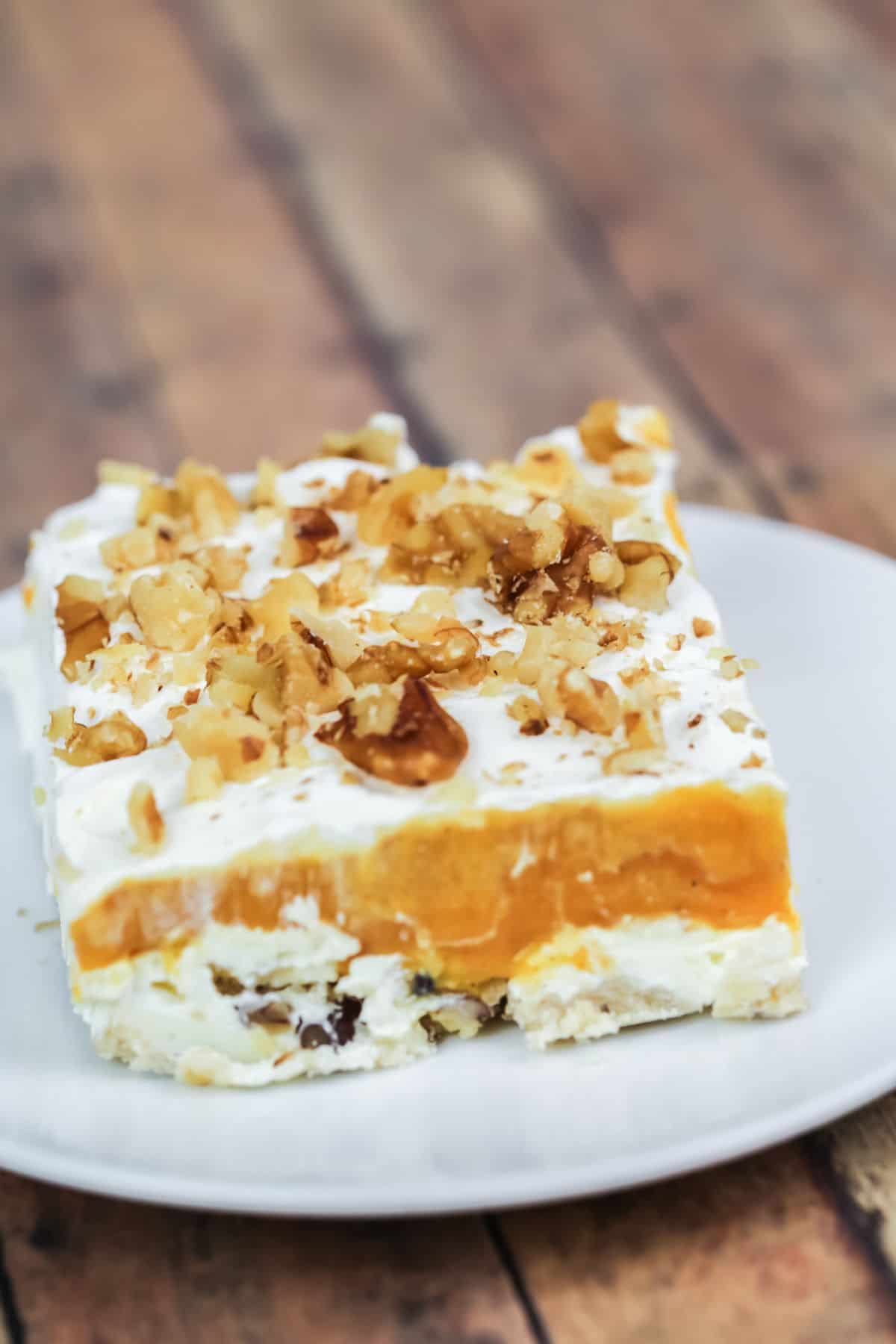 A square slice with a creamy white base, orange middle layer, whipped topping, and chopped walnuts, served on a white plate.