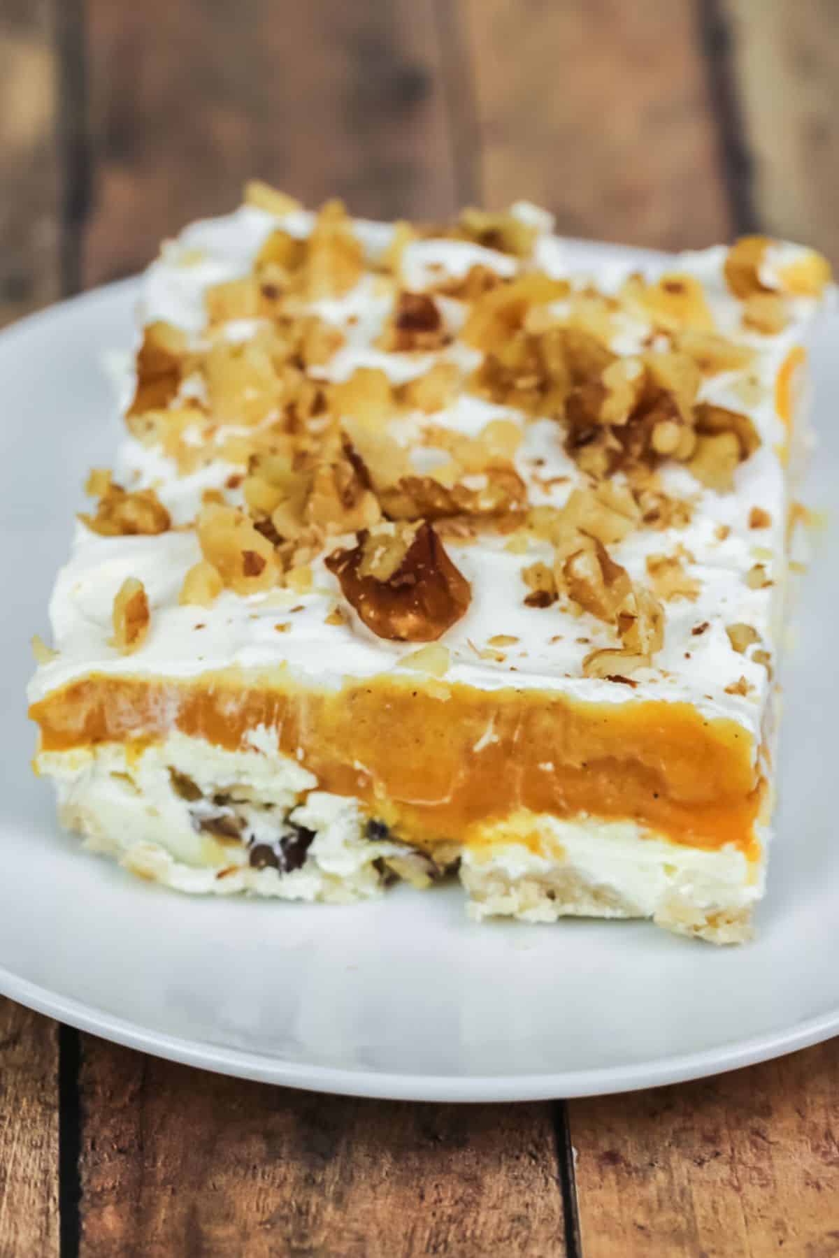 A square slice of Lush Cake featuring a creamy white base, bright orange layer, whipped topping, and chopped nuts is served on a white plate.