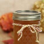 A glass jar filled with brown Pumpkin Pie Moonshine, sealed with a metal lid and decorated with a multicolored string bow, set against a blurred autumn-themed background.