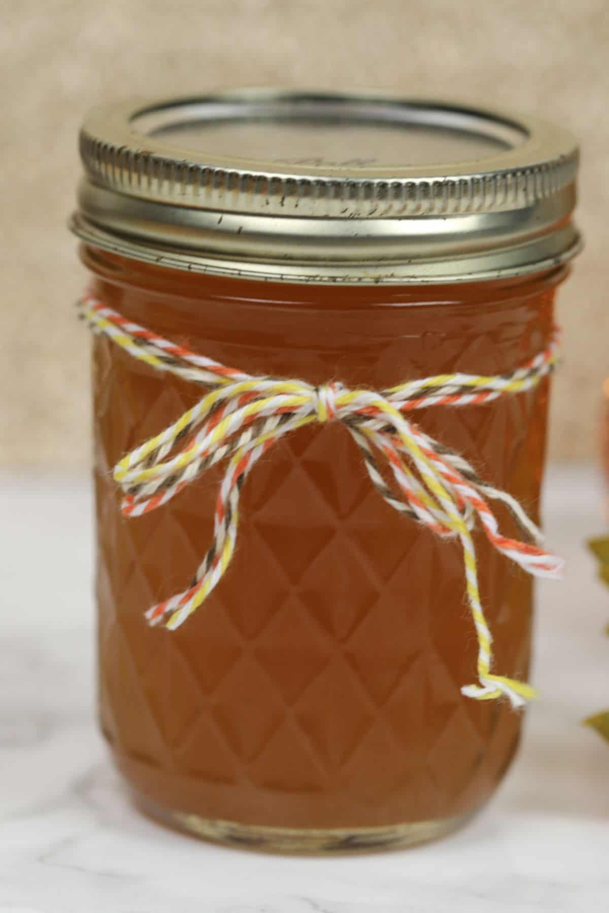 A glass jar filled with amber-colored Pumpkin Pie Moonshine, sealed with a metal lid and decorated with a red, yellow, and white twine bow.
