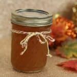 A glass jar filled with amber-colored drink sealed with a metal lid and adorned with a twine bow; autumn leaves and festive decorations set the cozy scene in the background.