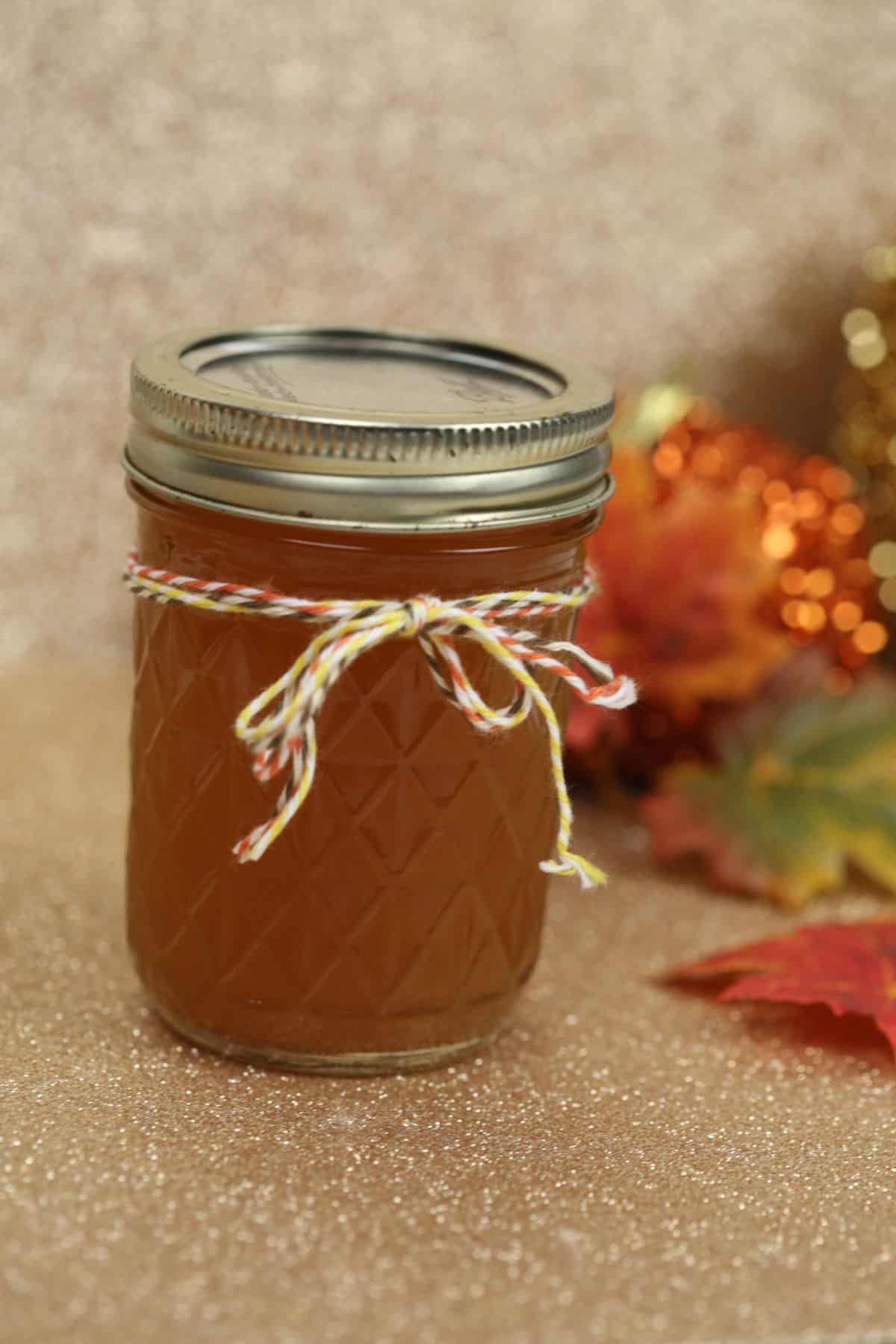 A glass jar filled with amber-colored drink sealed with a metal lid and adorned with a twine bow; autumn leaves and festive decorations set the cozy scene in the background.