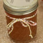 A glass jar filled with amber Pumpkin Pie Moonshine, sealed with a metal lid and decorated with a red, white, and yellow string bow, sits on a glittery gold surface.