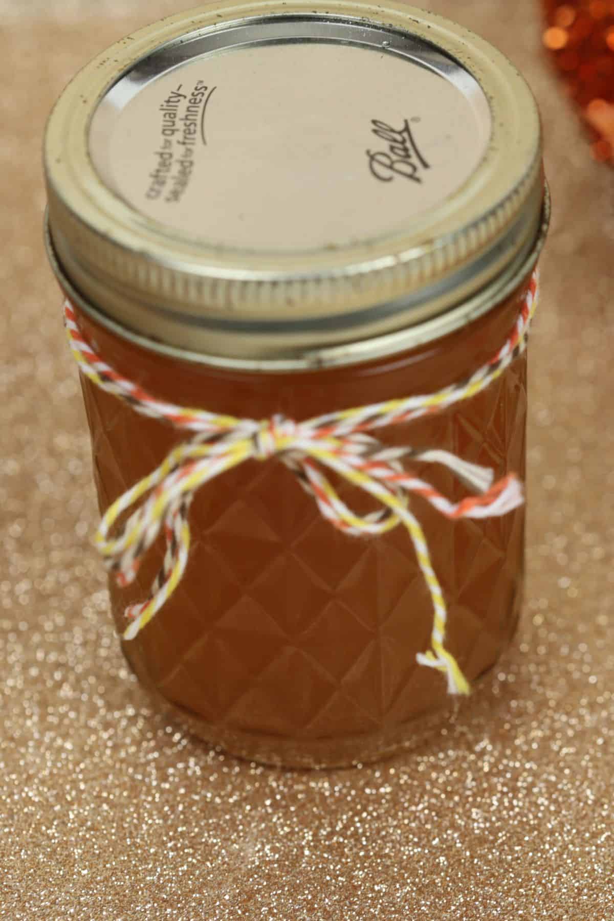 A glass jar filled with amber Pumpkin Pie Moonshine, sealed with a metal lid and decorated with a red, white, and yellow string bow, sits on a glittery gold surface.