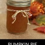 A glass jar of Pumpkin Pie Moonshine with a twine bow sits on a glittery surface, surrounded by autumn leaves and cozy decorations in the background.