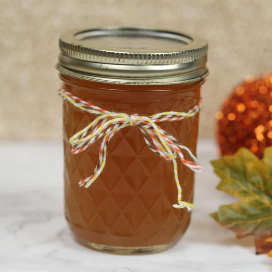 A glass jar filled with orange-colored Pumpkin Pie Moonshine, sealed with a metal lid and decorated with a multicolored string tied in a bow; an autumn leaf and decorative object in the background.