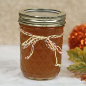 A glass jar filled with orange-colored Pumpkin Pie Moonshine, sealed with a metal lid and decorated with a multicolored string tied in a bow; an autumn leaf and decorative object in the background.