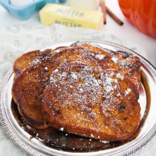 Slices of Pumpkin Pie Spice French Toast dusted with powdered sugar on a silver platter, with eggs, a stick of butter, a bottle of milk, and an orange pot in the background.