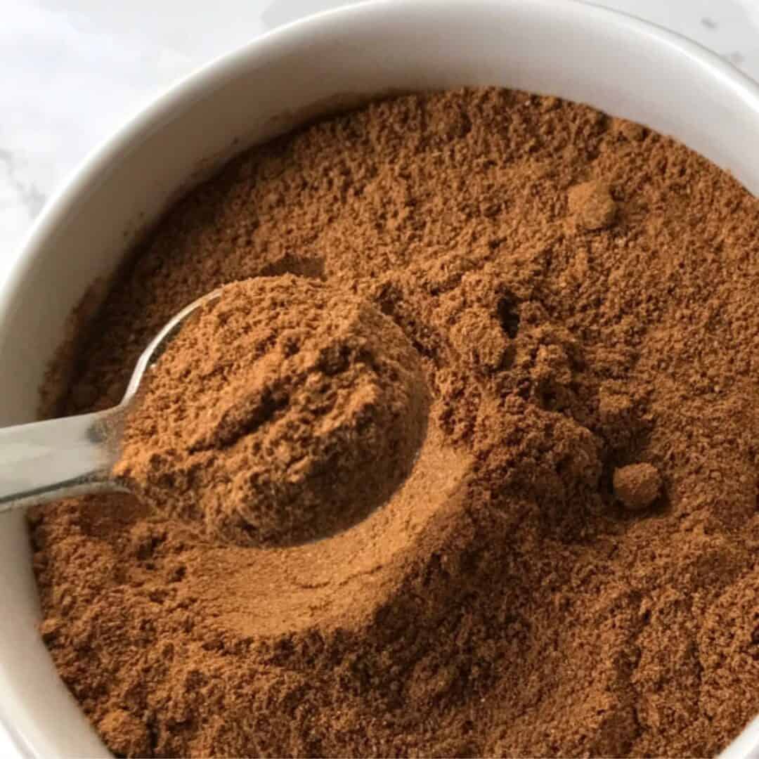 A metal spoon scoops ground cinnamon, a common Pumpkin Pie Spice Substitute, from a white bowl filled with more cinnamon powder.