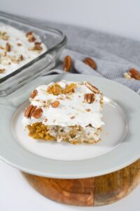 A slice of frosted Pumpkin Poke Cake with pecans sits on a white plate, while more of the cake rests in a glass dish, set against a gray cloth backdrop.
