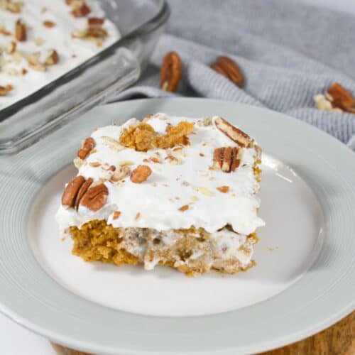 A slice of frosted Pumpkin Poke Cake with pecans sits on a white plate, while more of the cake rests in a glass dish, set against a gray cloth backdrop.
