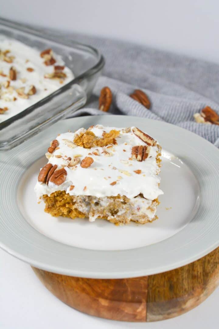 A slice of frosted Pumpkin Poke Cake with pecans sits on a white plate, while more of the cake rests in a glass dish, set against a gray cloth backdrop.