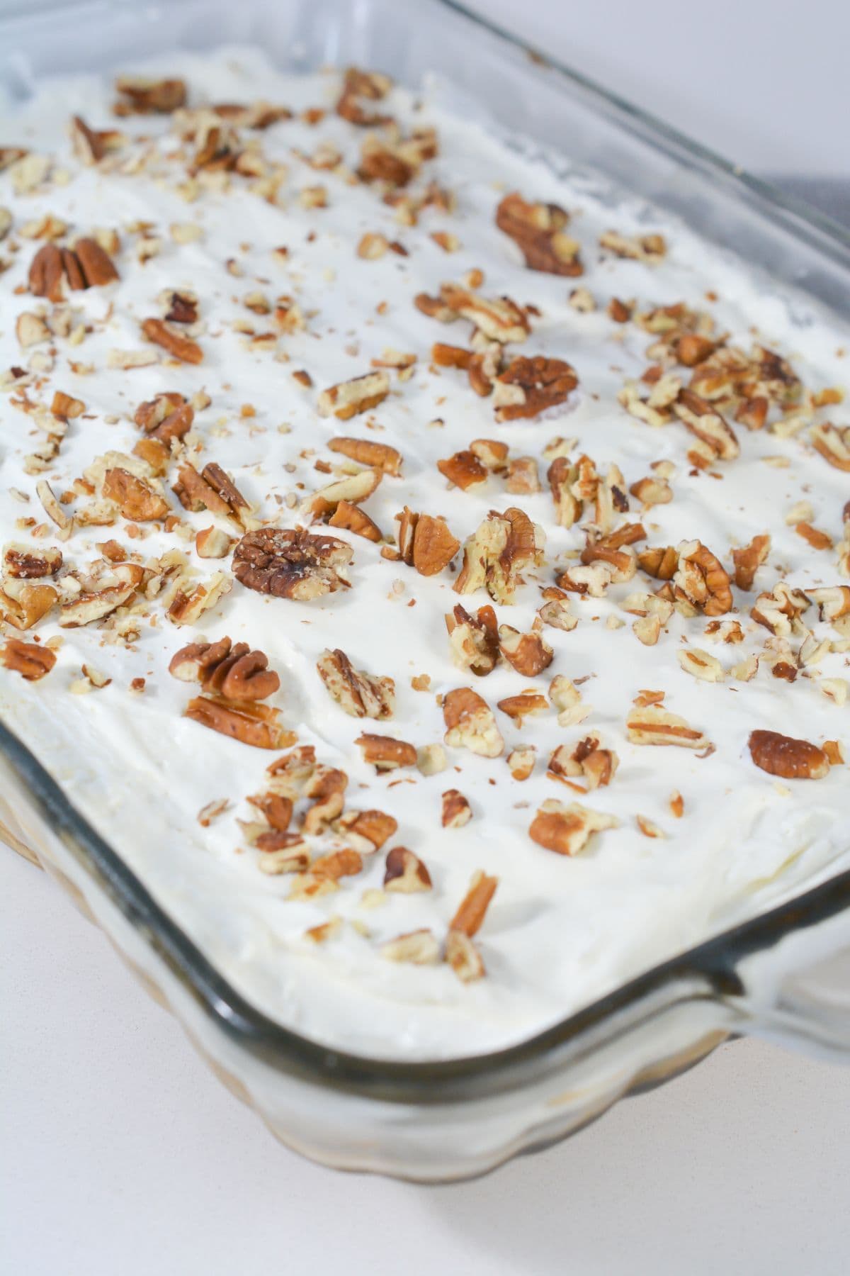A glass baking dish filled with dessert topped with creamy white frosting and a generous layer of chopped pecans.