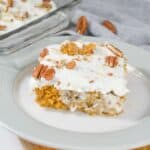 A slice of Pumpkin Poke Cake with white frosting and pecans on a white plate, with a pan of the same cake in the background.