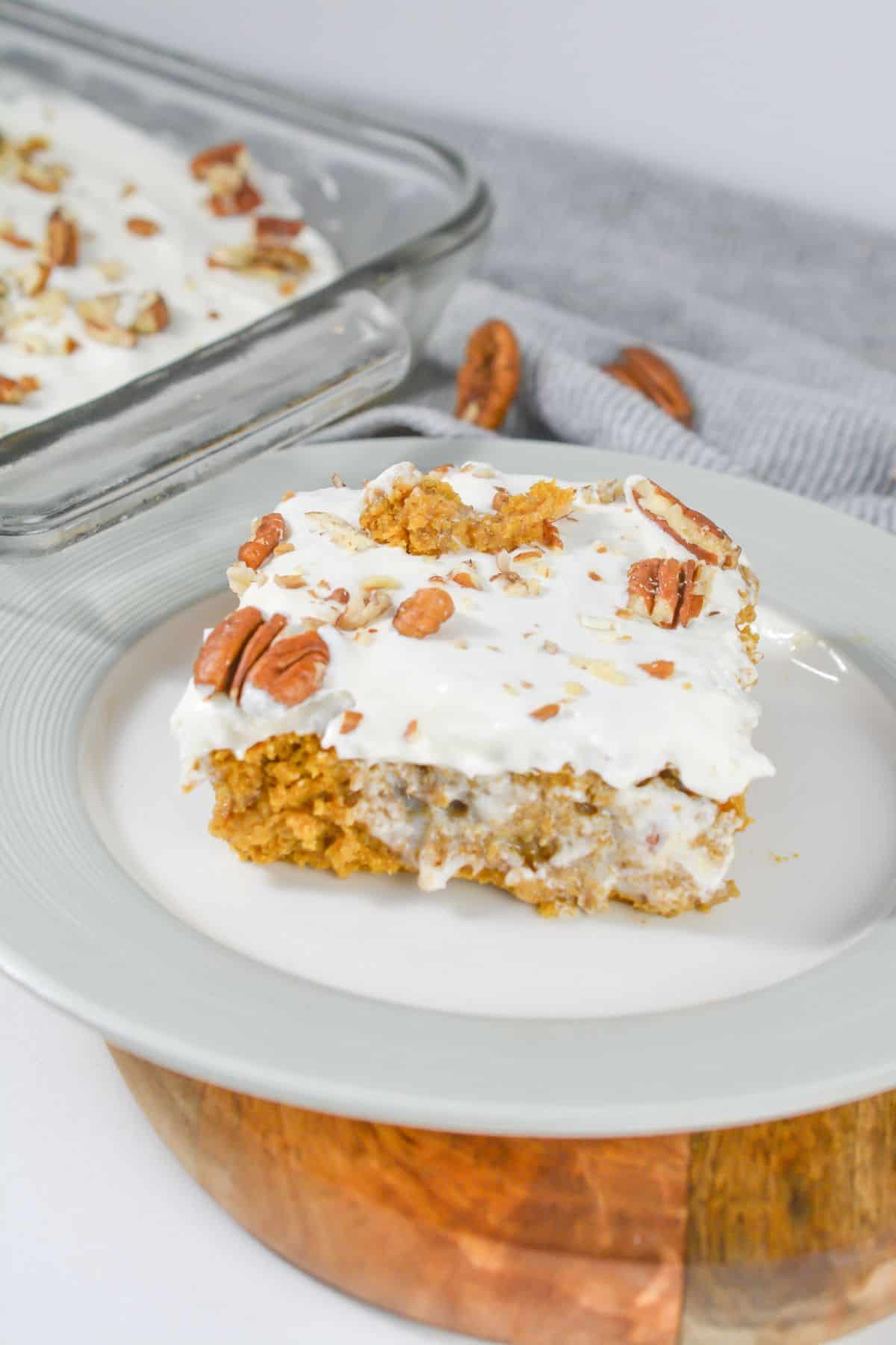 A slice of Pumpkin Poke Cake with white frosting and pecans on a white plate, with a pan of the same cake in the background.