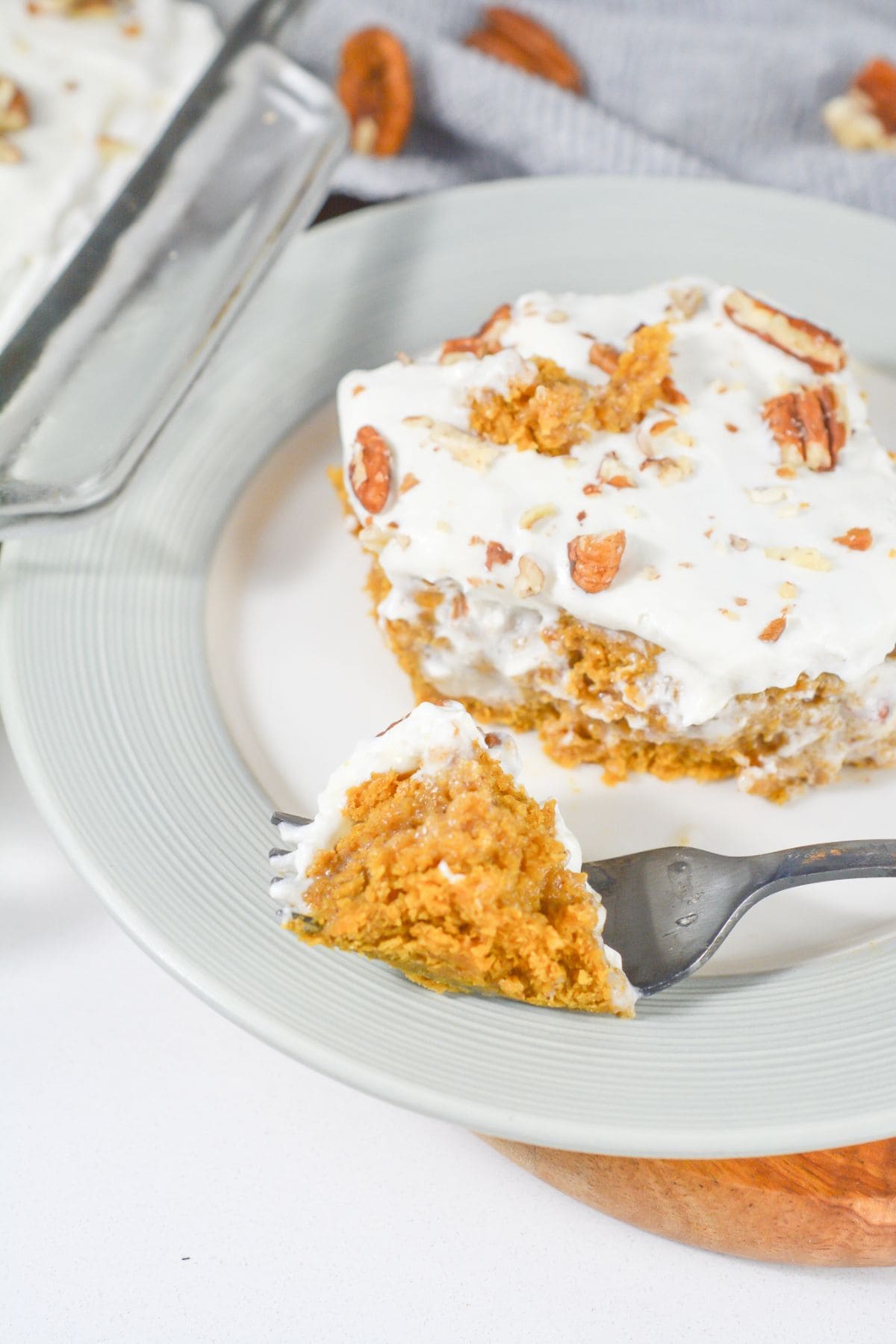 A slice of creamy Pumpkin Poke Cake topped with whipped topping and pecans sits on a plate, with a fork holding a bite in the foreground.