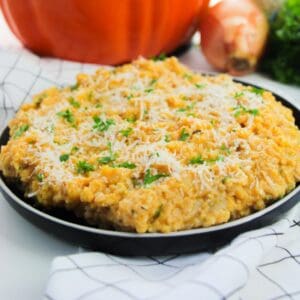 A plate of creamy pumpkin risotto topped with grated cheese and chopped herbs, set on a white surface with an onion and a pumpkin in the background.