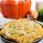 A plate of creamy Pumpkin Risotto topped with grated cheese and herbs sits in front of an orange pumpkin-shaped dish, with an onion and greens in the background.