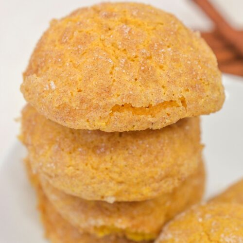 A stack of four round, golden-brown Pumpkin Snickerdoodle Cookies with a sugar-dusted, cracked surface sits on a white plate.