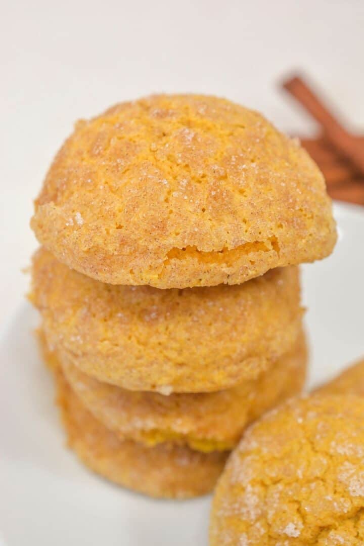 A stack of four round, golden-brown Pumpkin Snickerdoodle Cookies with a sugar-dusted, cracked surface sits on a white plate.