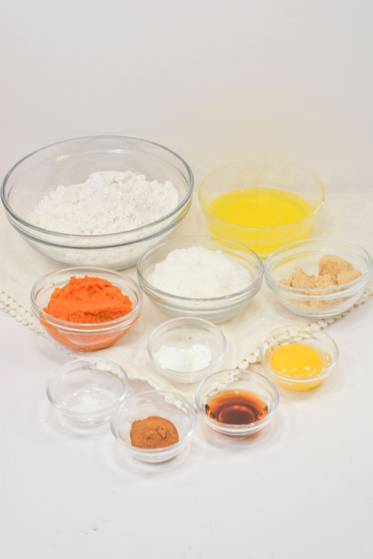 Assorted baking ingredients in glass bowls for Pumpkin Snickerdoodle Cookies, including flour, melted butter, pumpkin puree, sugar, egg yolk, vanilla extract, cinnamon, and baking powder, arranged on a white surface.