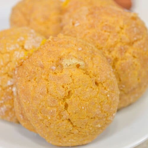 Three golden Pumpkin Snickerdoodle Cookies with a crackled surface rest on a white plate, with two cinnamon sticks in the background.