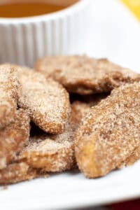 A close-up of cinnamon sugar-coated air fried apple slices on a plate, with a white cup partially visible in the background.