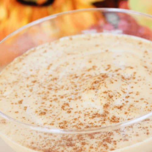 A Pumpkin Spice White Russian, this creamy dessert in a glass is topped with a sprinkle of cinnamon, perfectly complementing the blurred autumn-themed decorations in the background.