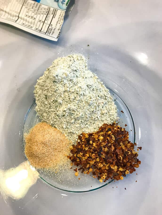Three piles of spices in a glass bowl: a mound of dried herb mix, a pile of granulated garlic, and a pile of red pepper flakes, perfect for creating ranch spicy crackers. Packaging with a nutrition label is partially visible in the background.