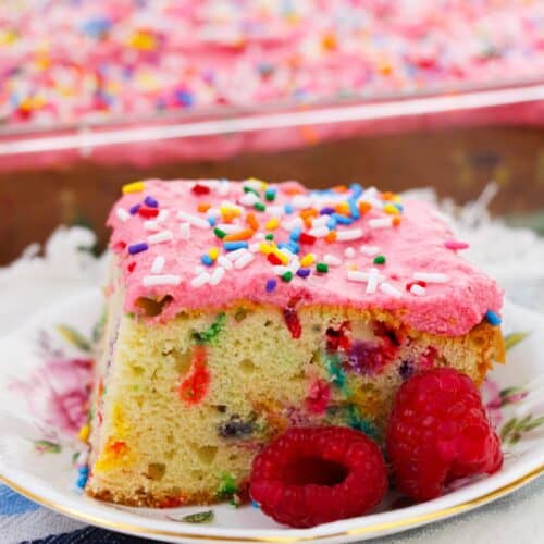 A slice of cake with pink raspberry buttercream frosting and colorful sprinkles sits on a floral plate with fresh raspberries, with the rest of the cake visible in the background.